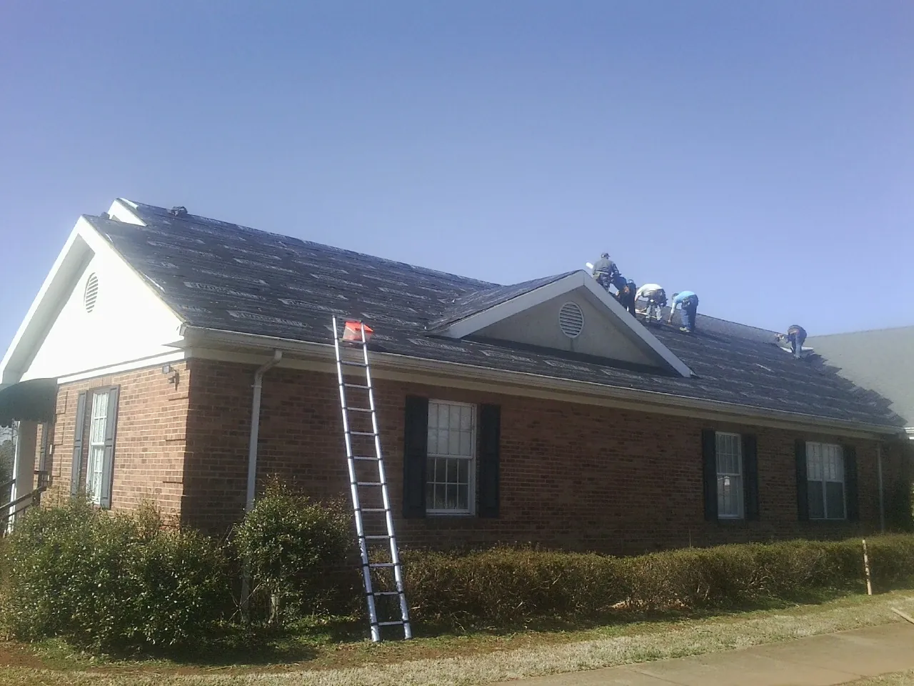 Crew tearing shingles off a brick ranch roof with a ladder set — Emergency Roof Repair work by our crew in Spurgeon