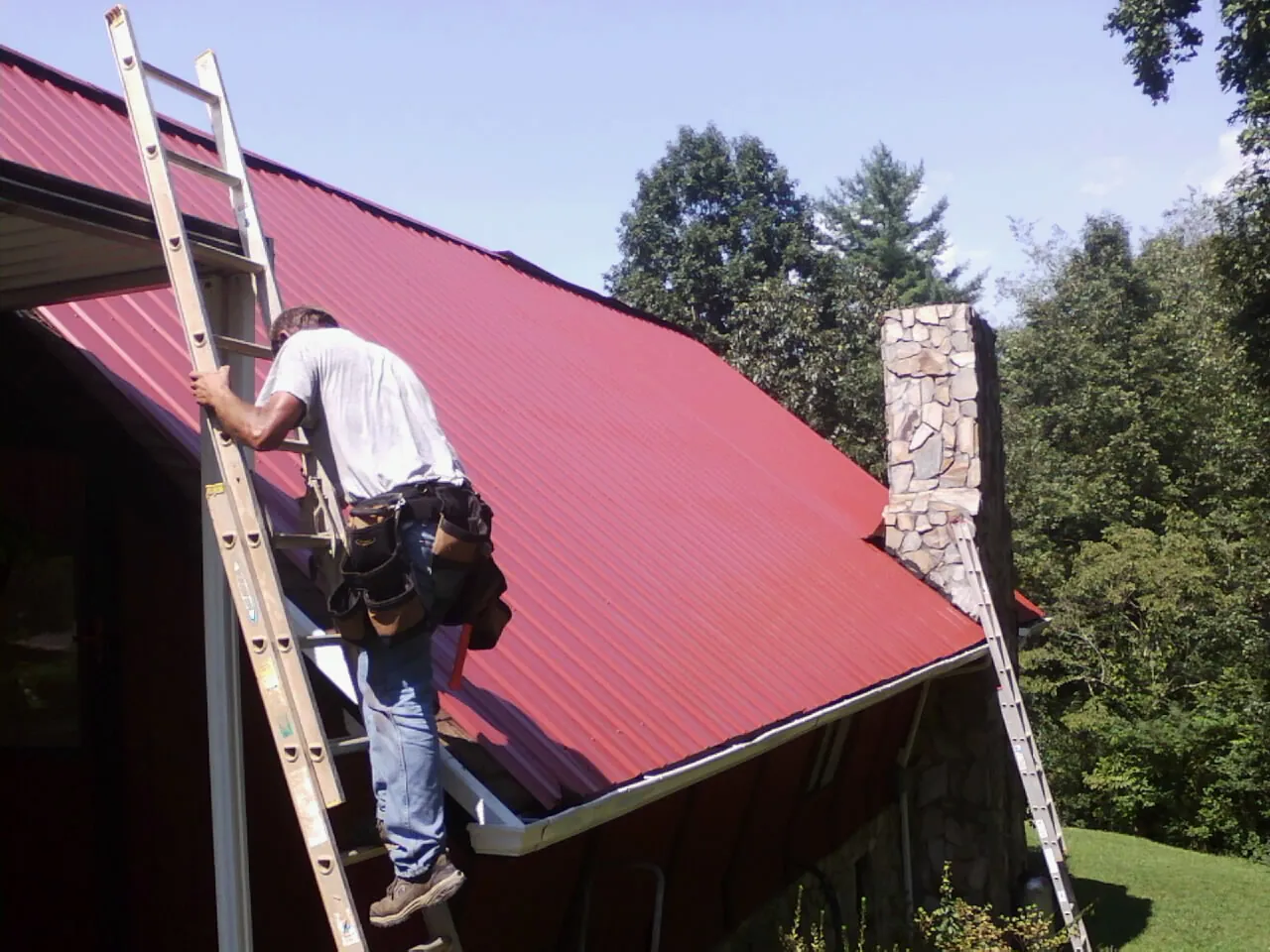 Roofer on a ladder beside a red metal roof and stone chimney — Soffit & Fascia Repair by Roofing Brothers in Spurgeon